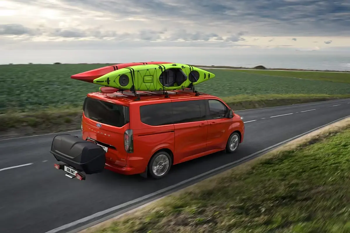 Ford Tourneo Custom - Ansicht bei der Fahrt, ein roter Tourneo Custom mit Sufbrettern auf dem Gepäckträger - Ford im Autocenter Mothor in Stendal, Gardelegen und Brandenburg kaufen, leasen oder finanzieren.